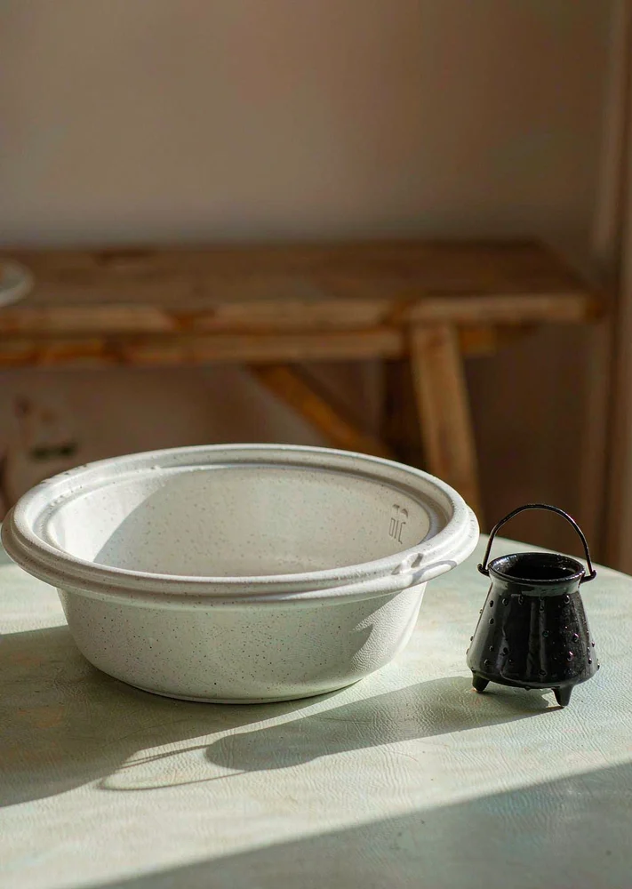 Wide white ceramic serving bowl beside a small black novelty cauldron on a light tabletop