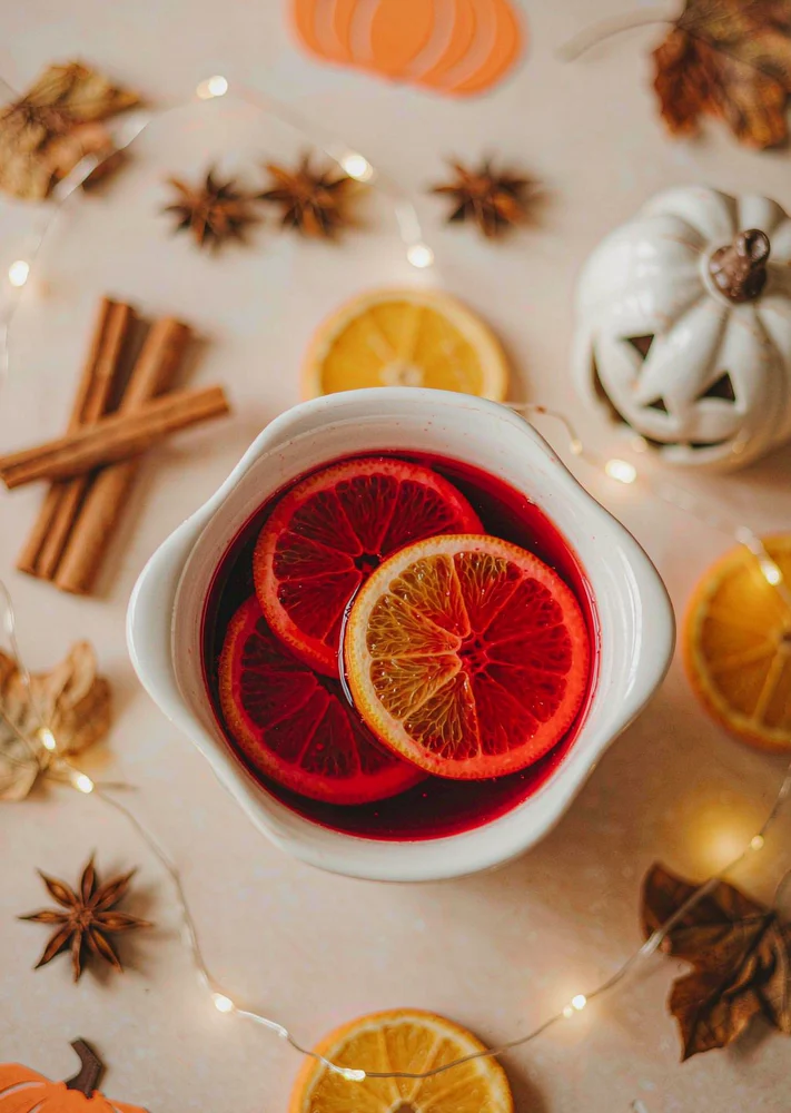 White ceramic bowl of red punch with orange slices, cinnamon sticks and warm fairy lights