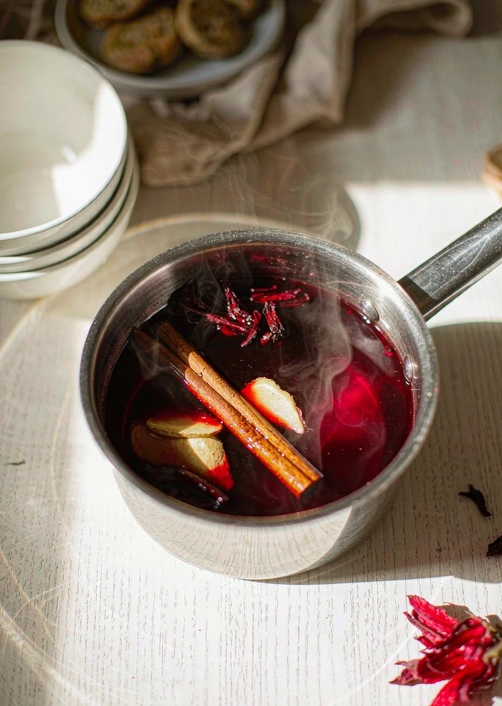 Stainless saucepan of ruby hibiscus tea with a cinnamon stick and fresh ginger on a light tabletop