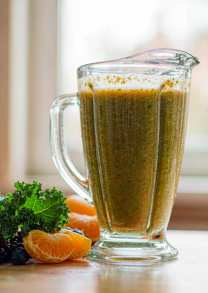 Clear glass blender jar of a green-orange kale, berry and orange smoothie on a light tabletop