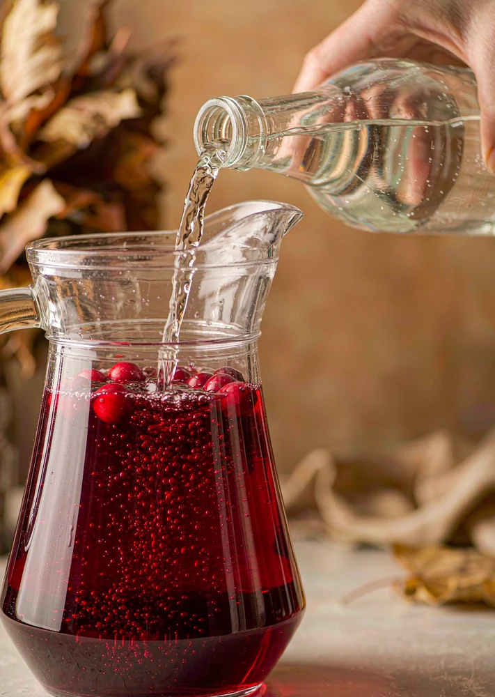 Glass pitcher of red cranberry punch with sparkling water being poured in, bubbles rising