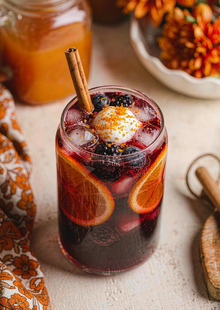 Tall clear glass of deep-red punch with orange slices, berries, a cinnamon stick and ice