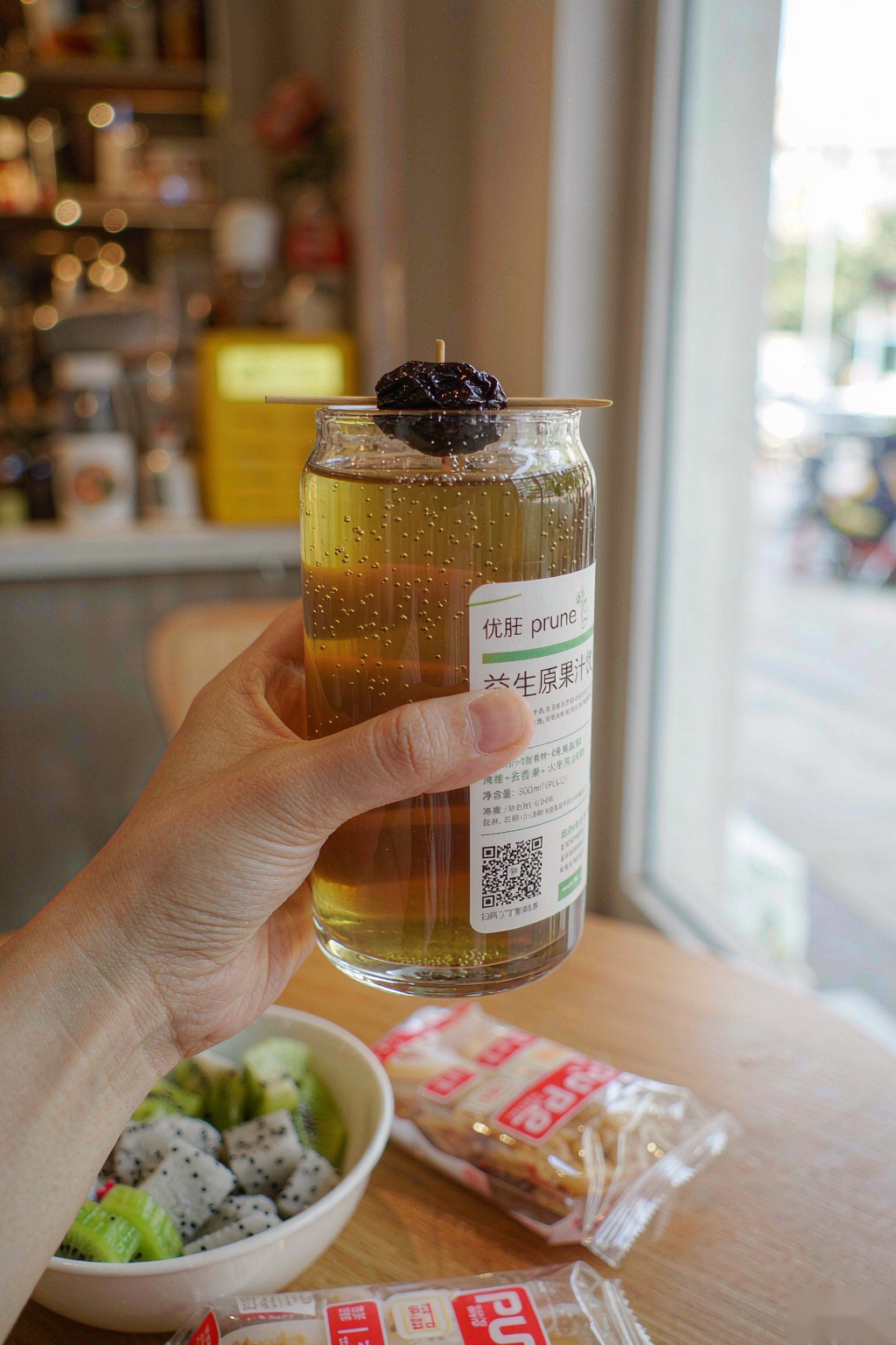 Hand holding a glass of Yougan Prune Detox Drink in a cozy cafe