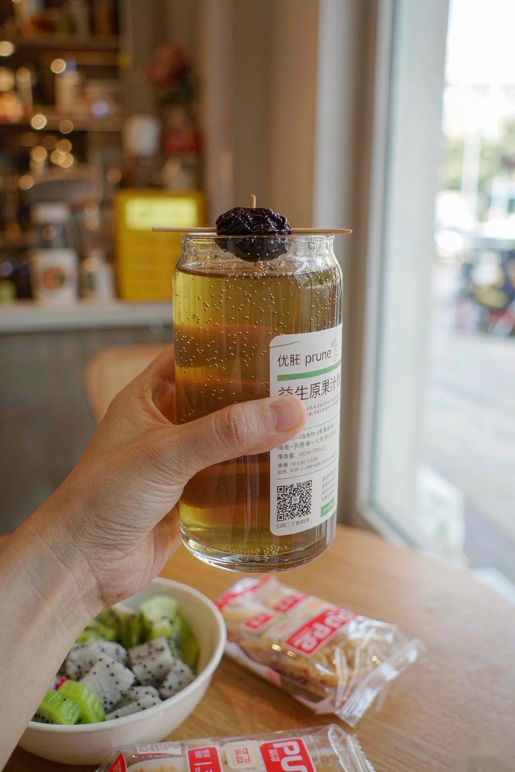 Hand holding a glass of Yougan Prune refresh Drink in a cozy cafe